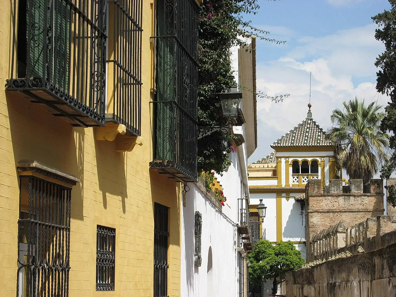 Sunny lane in Barrio Santa Cruz with yellow walls, iron window grilles and a tiled pavilion roof beyond, Seville