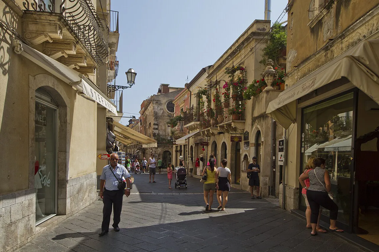 Sunlit Corso Umberto in Taormina lined with stone buildings, balconies and shop awnings