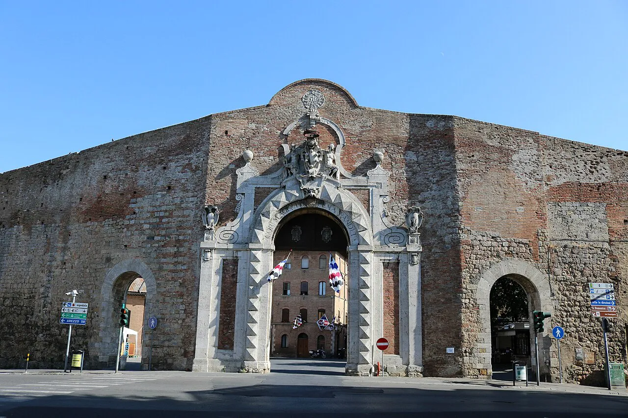 The carved stone archway of Porta Camollia set in Siena's brick city walls under a blue sky