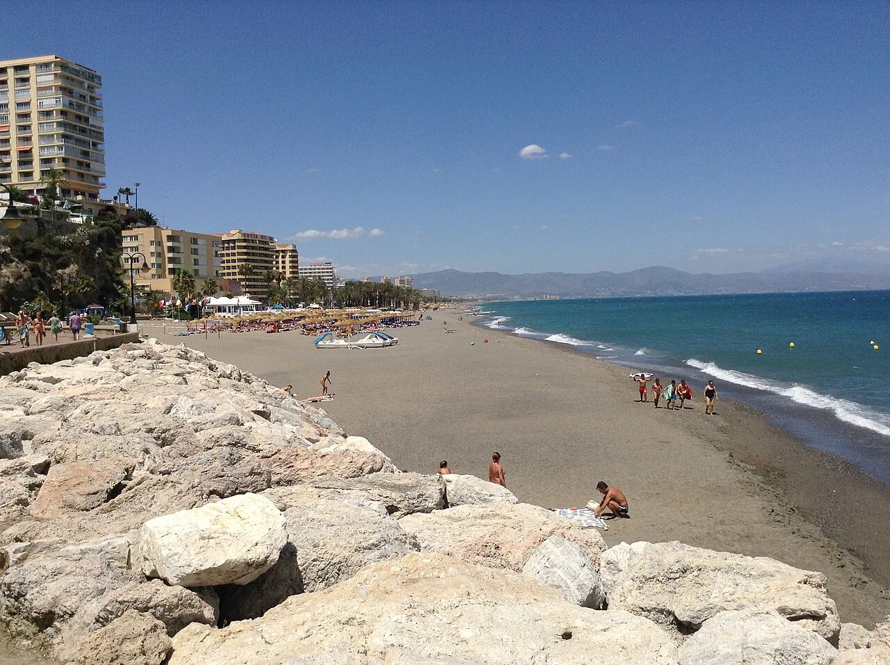 A rocky breakwater and long sandy beach on the Carihuela coast of Torremolinos under a blue sky
